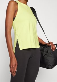 Limetkově zelený sportovní top s bočními rozparky, s malým bílým logem. Kombinován s černými legíny a černou sportovní taškou.