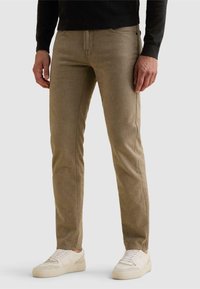 Homme portant un pantalon beige slim et des baskets blanches, debout devant un fond clair uni.