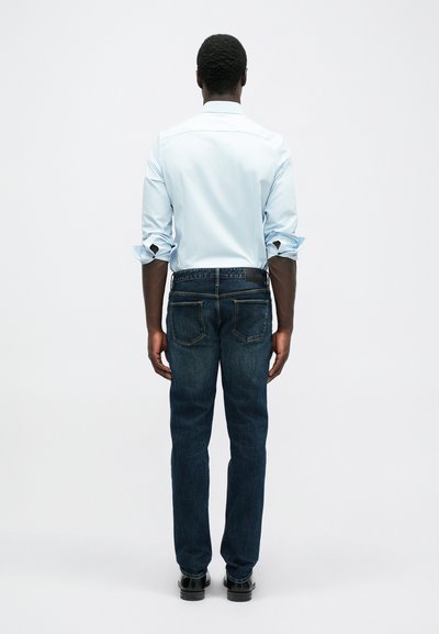 Ανοιχτό μπλε πουκάμισο με διπλωμένα μανίκια, συνδυασμένο με σκούρα μπλε τζιν που διαθέτουν πίσω τσέπες και μαύρη ζώνη. Μαύρα παπούτσια ολοκληρώνουν την εμφάνιση.