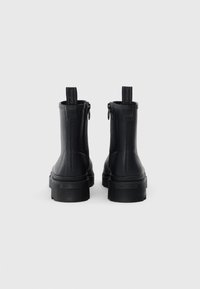 Une paire de bottines noires à chevilles avec des semelles épaisses et des languettes arrière, vues de l'arrière sur un fond clair.