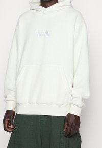 Weißer Fleece-Hoodie mit Kängurutasche, elastischen Bündchen und Saum sowie dezentem, auf die Brust gesticktem Logo. Weiche Textur und lockere Passform.