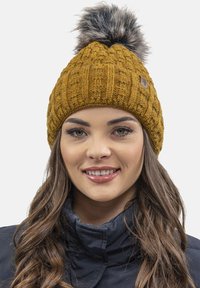 Gorro de punto color mostaza con un patrón texturizado, que presenta un pompón de piel sintética en la parte superior y una pequeña etiqueta de la marca en un lado.