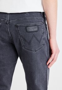 Tamsiai pilki džinsai su užpakaliniu kišenėle, pažymėta "Wrangler" logotipu ir išskirtiniu siuvinėjimo raštu. Lygi tekstūra, pritaikytas kirpimas.