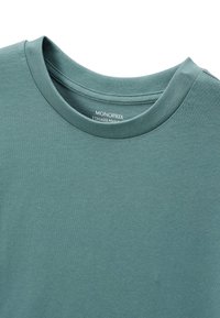 T-shirt en coton teal avec un col rond, présentant une texture douce et une étiquette à l'intérieur qui indique "MONOPRIX."