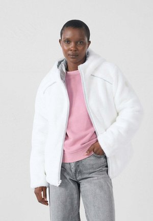 Vrouw met kort haar in een witte fleece jas met ritssluiting over een roze sweatshirt en grijze spijkerbroek, staand tegen een effen lichte achtergrond.