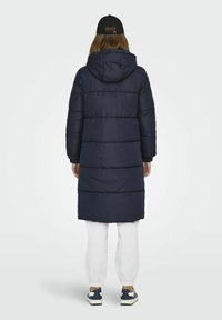 Doudoune marine avec capuche, présentant des sections matelassées, des manches longues et une longueur mi-mollet, assortie à un survêtement gris clair et des baskets.