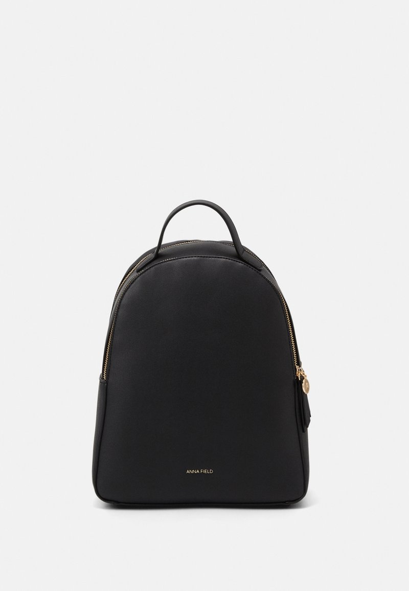 Sac à dos en cuir noir de forme arrondie, avec une fermeture éclair dorée et une petite poignée. Présente un marquage subtil en bas à l'avant. Design minimaliste.