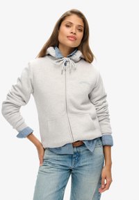 Sudadera con cremallera de color gris claro, con capucha ajustable, bolsillos frontales y branding bordado. Lleva puesta sobre una camisa de mezclilla y combina con jeans azules.