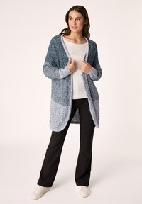 Cardigan lavorato a maglia in gradiente blu e grigio, con design aperto, trama leggera, maniche lunghe, abbinato a pantaloni neri e sneakers bianche.