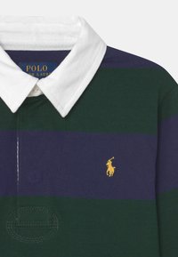 Polokošile s bílým límečkem, se zelenými a fialovými vodorovnými pruhy. Malé žluté logo koně na levé části hrudi. Bavlněný materiál.