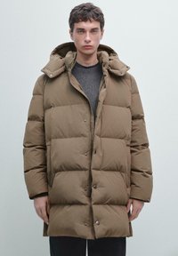 Cappotto imbottito marrone con cappuccio, dotato di chiusura con zip e bottoni a pressione, texture trapuntata e una vestibilità rilassata. Indossato sopra una maglia grigia.