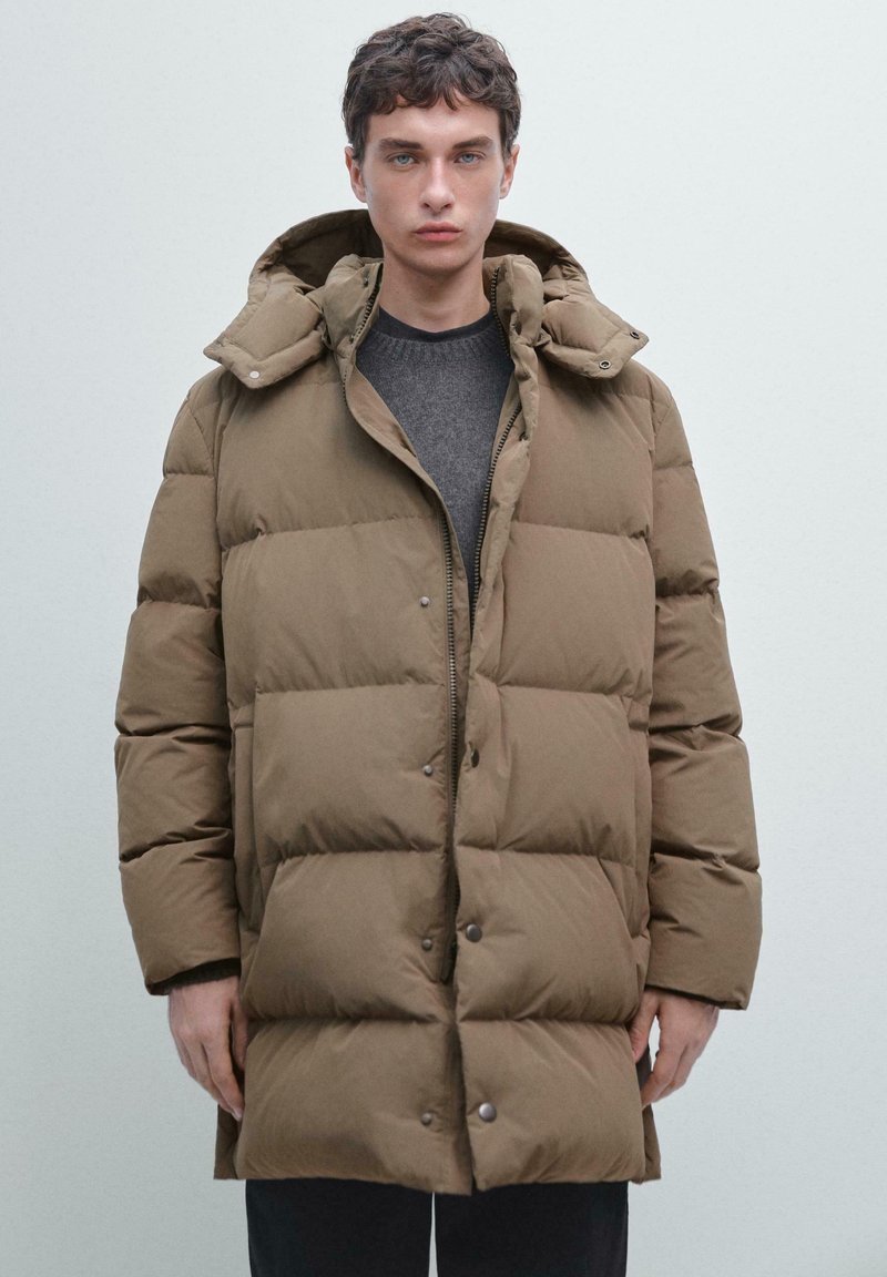 Cappotto imbottito marrone con cappuccio, dotato di chiusura con zip e bottoni a pressione, texture trapuntata e una vestibilità rilassata. Indossato sopra una maglia grigia.
