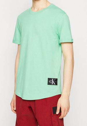T-shirt in cotone verde menta con collo rotondo, maniche arrotolate e un'etichetta nera con il logo "Calvin Klein Jeans" vicino all'orlo.