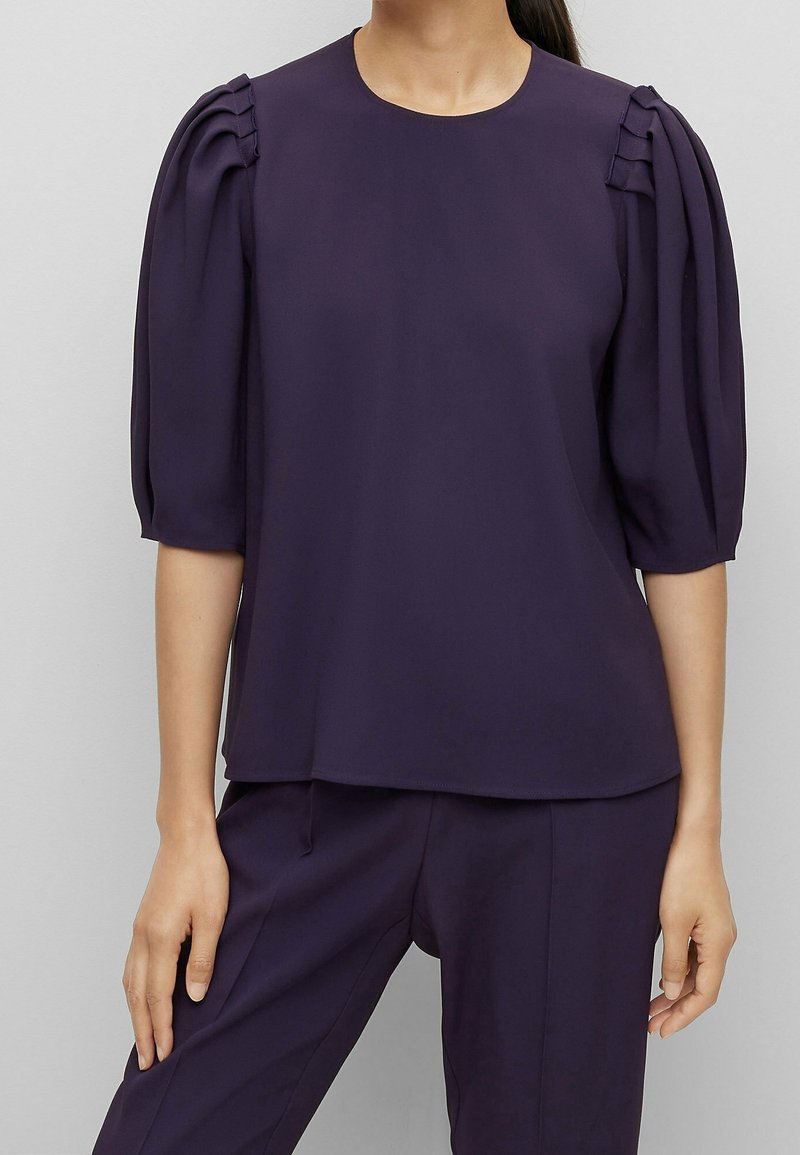 Femme portant un chemisier violet foncé avec des manches bouffantes au niveau du coude et un pantalon violet foncé assorti sur un fond uni.