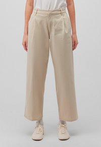 Beige weite Hosen mit Falten, aus einem glatten Stoff. Mit einem hohen Bund und seitlichen Taschen ausgestattet, kombiniert mit hellen Schuhen.