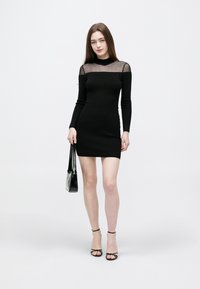 Černé mini šaty s žebrovanou texturou, dlouhými rukávy, průhledným výstřihem a zdobenými detaily. Kombinované s černými sandály na podpatku a malou kabelkou.