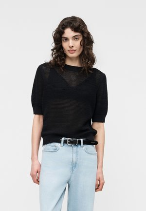 Nuori nainen, jolla on kiharat hiukset, pukeutuneena mustaan lyhythihaiseen neulepaitaan, joka on tungettu vaaleansinisiin farkkuihin mustan vyön kanssa.