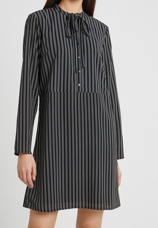 Robe à manches longues rayée noir et blanc avec un col à nouer, une patte de boutonnage et un tissu doux. Silhouette évasée avec une longueur mi-cuisse.