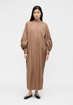 Nő áll egy barna, hosszú ujjú maxi ruhában, melynek felfújt ujjain virágos hímzés található, fekete lapos cipővel, fehér háttér előtt.