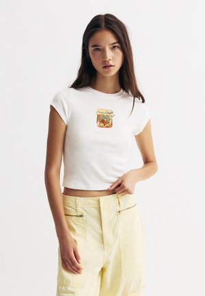 Hvid cropped t-shirt med korte ærmer, der har et illustreret glasdesign. Stylet med gule cargo-bukser og en neutral baggrund.