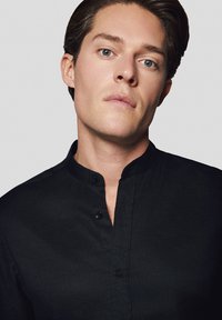 Czarna koszula z guzikami i kołnierzykiem mandaryńskim, z teksturowanej tkaniny i listwą guzikową z przodu. Zbliżenie na dekolt i guziki.