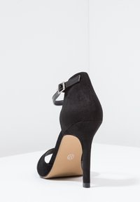 Musta saappaankorko sandaalit, joissa on nilkkaremmi, suljettu takaosa ja terävä kärki. Varustettu vaaleanruskealla pohjalla ja kapealla stiletto-korkealla.