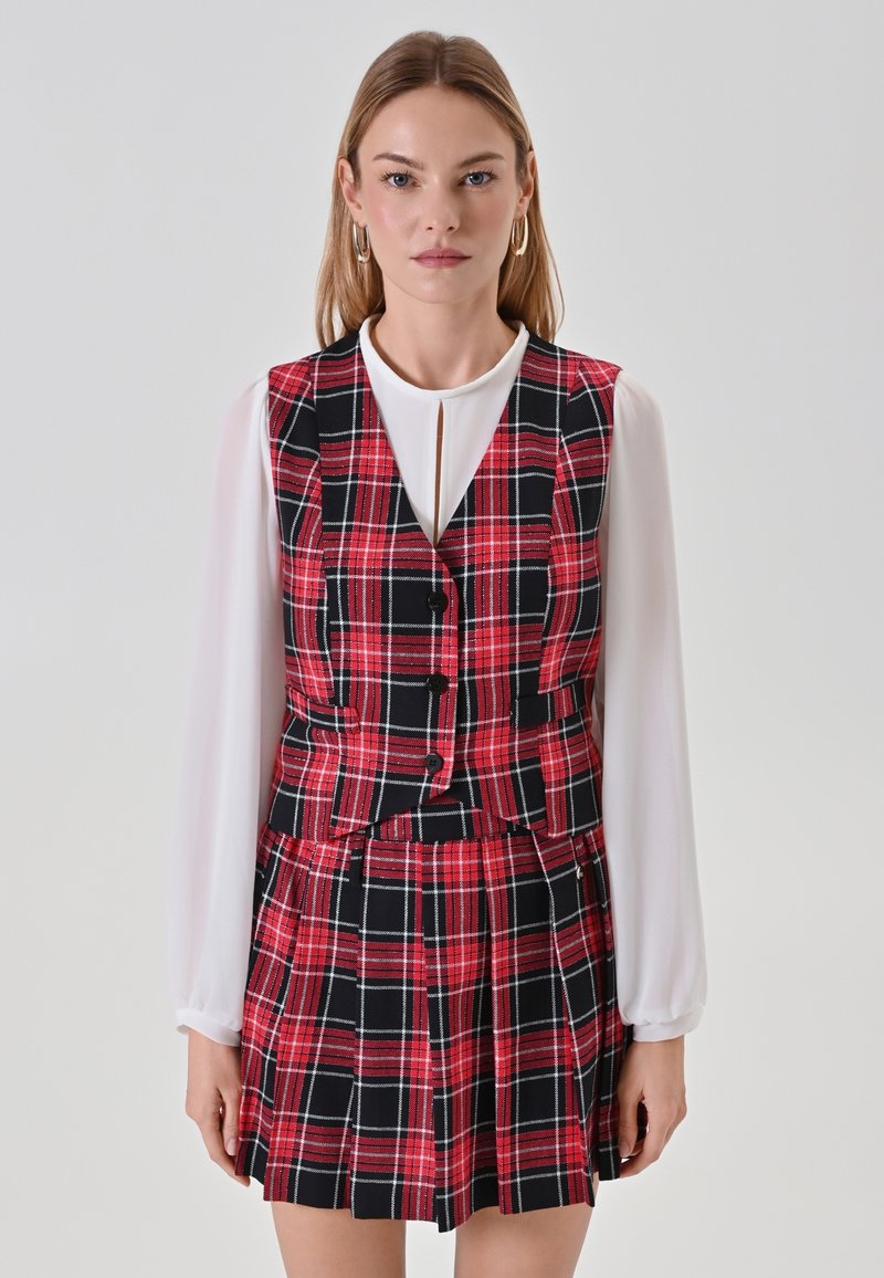 Completo in tessuto scozzese rosso e nero composto da gilet e gonna, abbinato a una camicia bianca a maniche lunghe. Include bottoni, pieghe e un colletto.