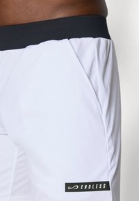 Nærbilde av hvite idrettsshorts med svart livbelt og en svart etikett med teksten "ENDLESS" på høyre side nær kanten.