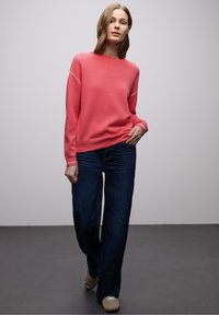 Femme portant un pull corail à manches longues, un jean large foncé et des baskets beige, debout devant un fond gris uni.