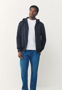 Tmavomodrá mikina na zips s nastaviteľnými šnúrkami, na sebe biela tričko, v kombinácii s modrými džínsami; má mäkkú textúru a štandardný strih.