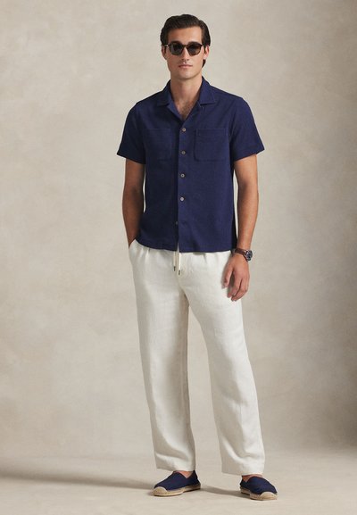 Άνδρας με σκούρο μπλε κοντομάνικο πουκάμισο με κουμπιά, λευκό παντελόνι με κορδόνι, ναυτικά εσπαντρίγιες, γυαλιά ηλίου και ρολόι χειρός, στέκεται μπροστά σε απλό φόντο.