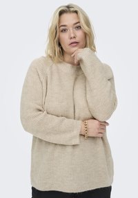 Beige Strickpullover mit rundem Ausschnitt, gerippten Bündchen und Saum sowie einer lockeren Passform; getragen mit einem goldenen Kettenarmband am Handgelenk.