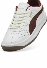 Białe sneakersy z brązowymi akcentami, wykonane ze skóry. Posiadają perforowaną wkładkę na palce, płaskie sznurowadła, wyściełany kołnierz oraz gumową podeszwę.