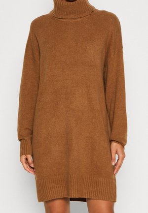 Femme portant une longue robe-pull à col roulé en maille marron avec des poignets et un ourlet côtelés, debout devant un fond uni.