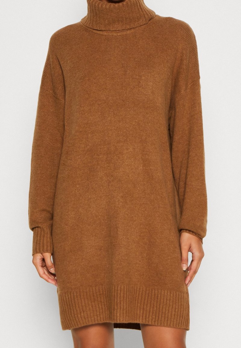 Femme portant une longue robe-pull à col roulé en maille marron avec des poignets et un ourlet côtelés, debout devant un fond uni.