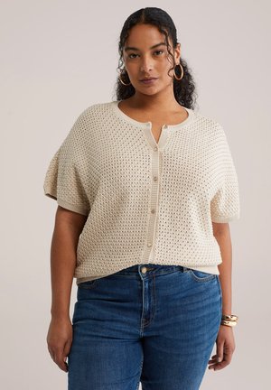 Béžový pletený sveter s krátkymi rukávmi, so zapínaním na gombíky vpredu a textúrovaným vzorom. Kombinovaný s modrými džínsami z denimu.