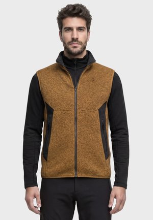Mann trägt eine braune ärmellose Fleeceweste mit Reißverschluss über einem schwarzen Langarmshirt und steht vor einem schlichten hellen Hintergrund.