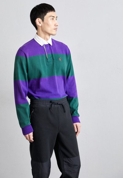 Polo de manga longa às riscas roxas e verdes com gola, logótipo no peitoral; combinado com calças pretas com cordão de ajuste e zíperes laterais.