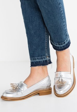Pieds portant des mocassins en cuir argenté avec des glands, associés à un jean denim court bleu foncé avec des ourlets effilochés sur fond blanc.