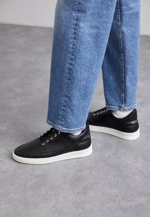 Schwarze Ledersneaker mit weißen Gummisohlen, ausgestattet mit runden Schnürsenkeln, glatter Oberfläche und dezentem Branding an der Ferse. Getragen mit blauen Jeans.