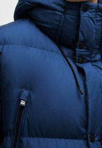 Blue padded jacket with a high neck and hood, featuring black hardware, buttons, and a zippered side pocket. Smooth, textured fabric.