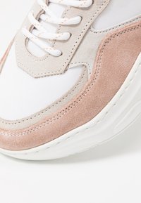 Gros plan d’une basket blanche avec des empiècements en suède beige et rose, des lacets blancs avec des coutures orange et une semelle blanche épaisse.
