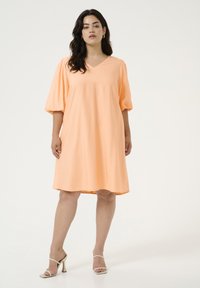 Robe en coton pêche avec un décolleté en V, des manches courtes bouffantes et une coupe ample, se terminant au-dessus des genoux. Accordée avec des sandales à talons beige.