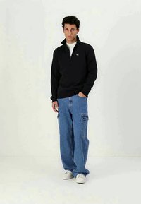 Black zip-neck sweatshirt, loose blue cargo jeans with pockets, and white sneakers. Model standing against a plain background.