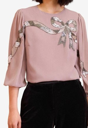 Mauve bluse med puffede trekvartærmer, sort og sølv palietbånd og sløjfe pynt tværs over brystet og ærmerne, kombineret med sorte bukser.