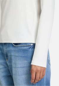 Langärmliges weißes Rippoberteil, kombiniert mit hellblauen Denim-Jeans. Das Oberteil hat einen runden Ausschnitt und enganliegende Ärmel.