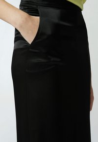 Crna satenska suknja s bočnom džepom, koja se odlikuje glatkom teksturom i elegantnim, prilagođenim dizajnom. Vidljive šavove i detalj visokog struka.