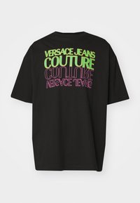 Svart oversized t-shirt med fet, färgglad text: "VERSACE JEANS COUTURE" i grönt och rosa, med en mjuk bomullstextur.