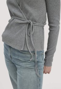 Grauer gerippter Wickeloberteil mit gebundenem Taillenverschluss, kombiniert mit hellblauen Jeans. Die Textur ist weich, und das Design hat lange Ärmel.