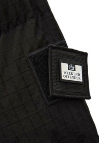 Schwarzer Stoff mit Textur, der ein Gittermuster zeigt, mit einem rechteckigen Patch, der in weißer und schwarzer Stickerei "WEEKEND OFFENDER" beschriftet ist.